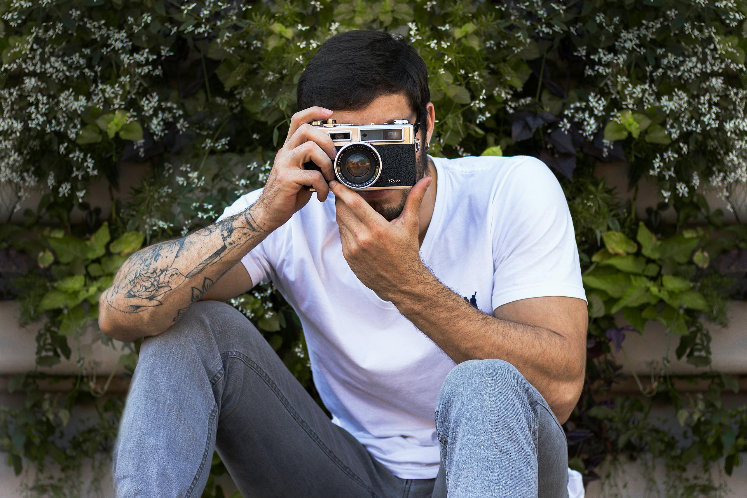 Anonymous tattooed male in stylish apparel taking photo on digital camera while sitting near blooming shrub