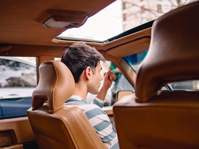 Back view of a young man sitting in a vintage car with a sunroof, enjoying a sunny day.