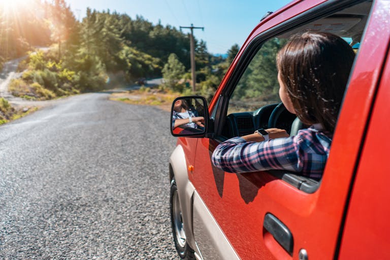 Back view of unrecognizable female driver looking out of window while riding red automobile on asphalt road near green trees