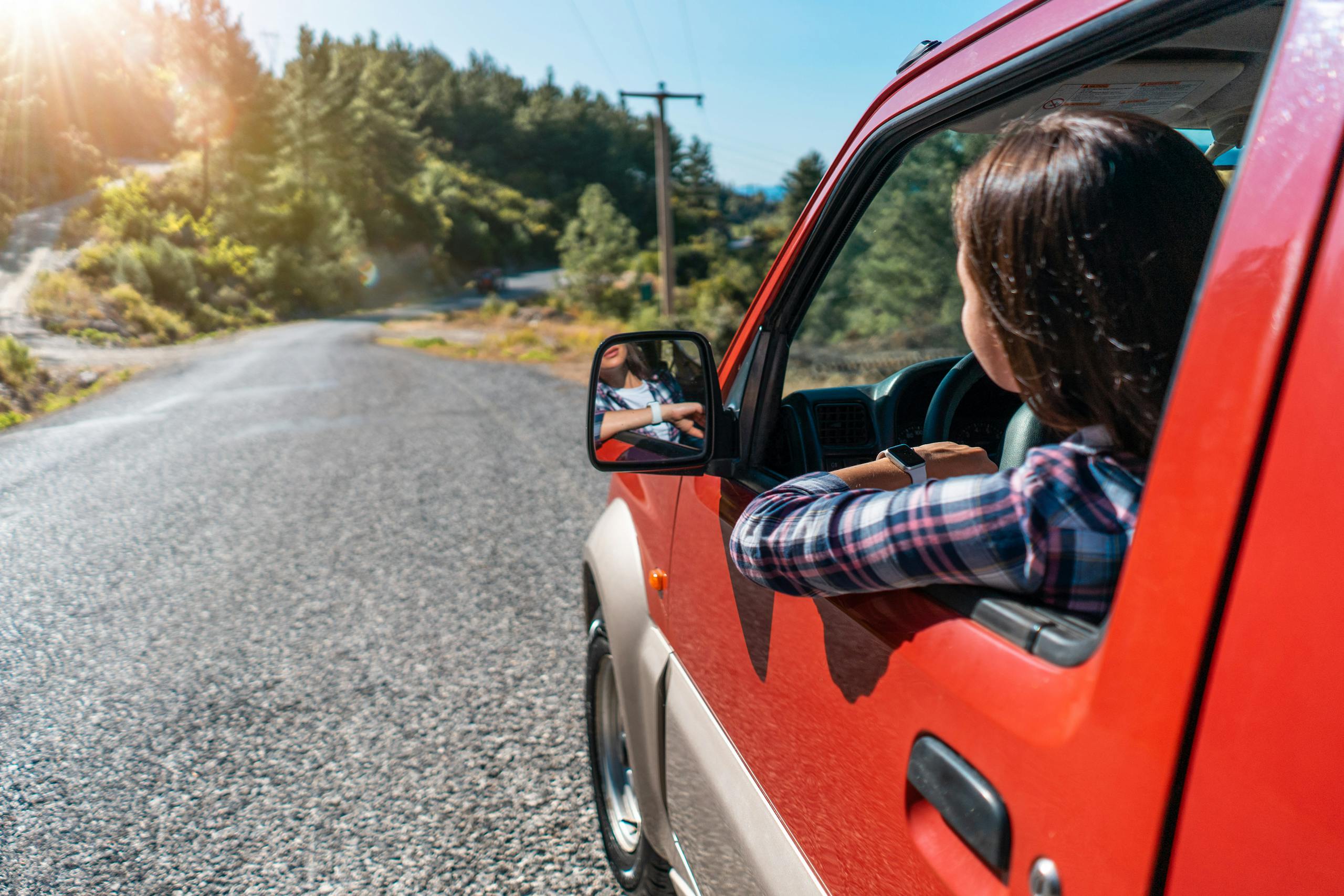 Back view of unrecognizable female driver looking out of window while riding red automobile on asphalt road near green trees