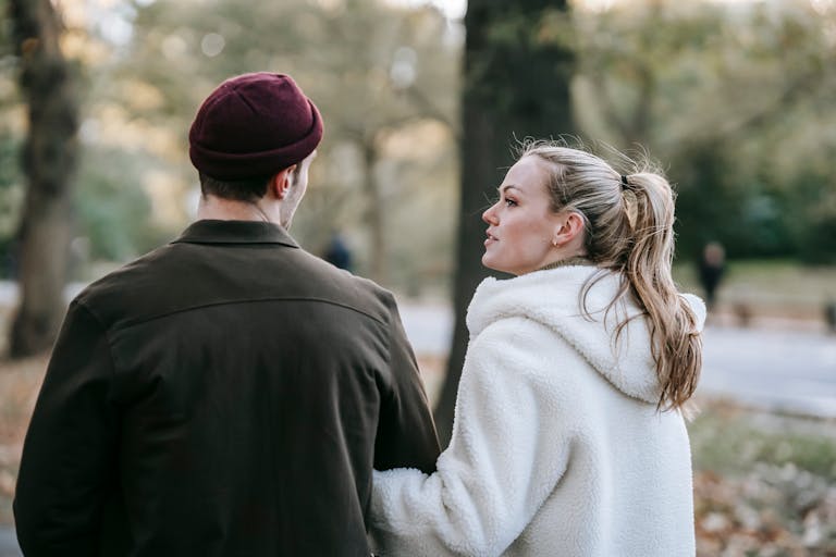 Back view of young stylish lady with blond hair in stylish warm outerwear holding hand of anonymous boyfriend while strolling together in autumn park