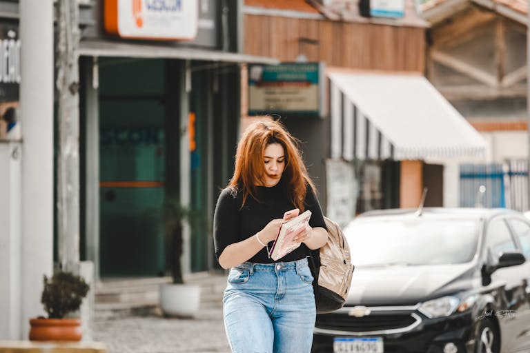 Casually dressed woman with red hair checking her smartphone on a busy city street.