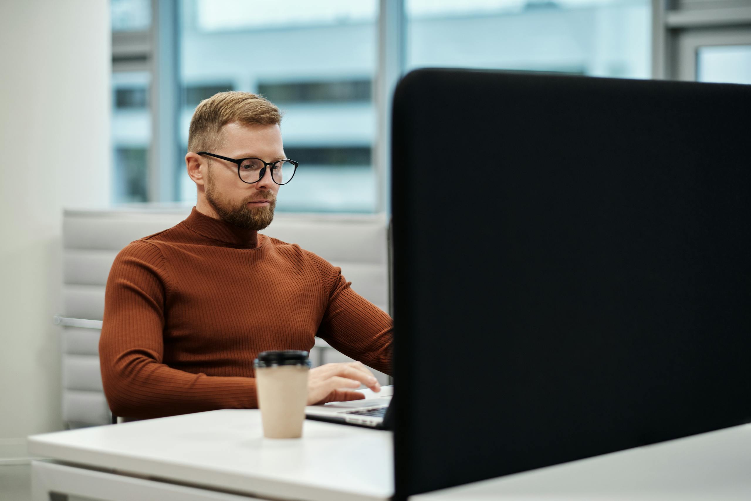 Caucasian man with beard and glasses working at a desk in a modern office environment.
