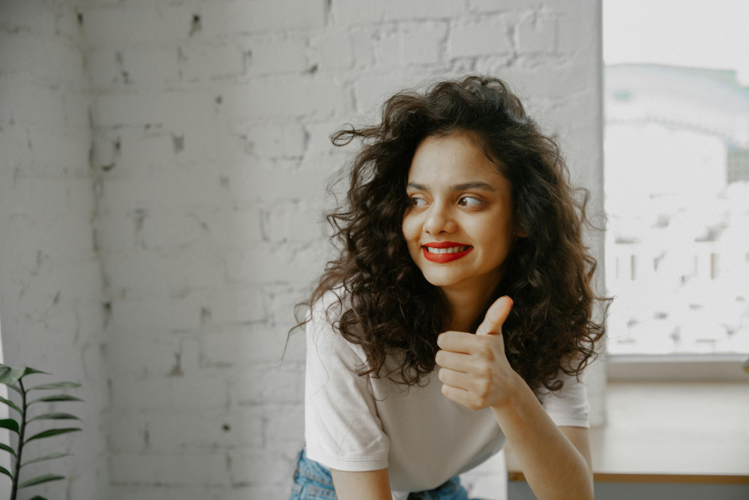 Cheerful woman with curly hair smiling and giving thumbs up gesture indoors.