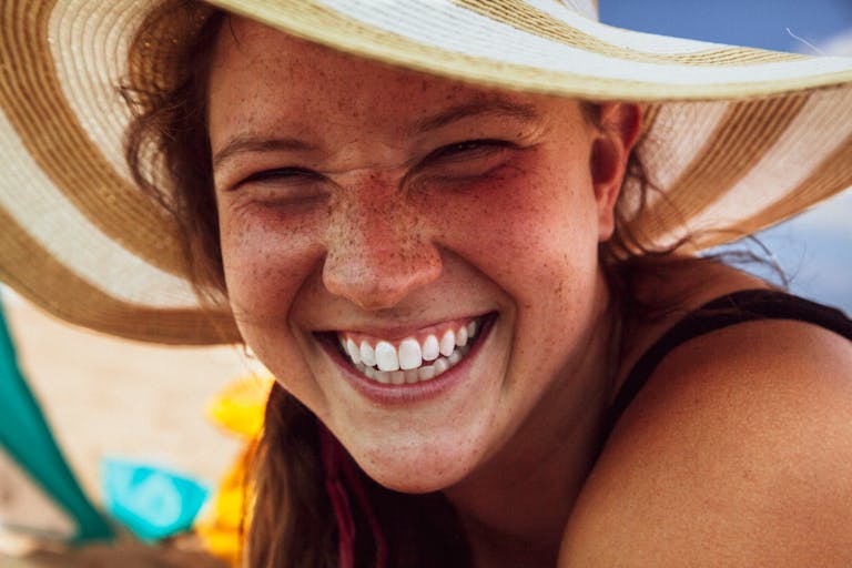 Close-up of a smiling woman with freckles wearing a sun hat, exuding happiness.