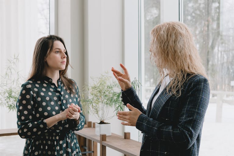 Concentrated young female colleagues in elegant outfits standing near window in light office and discussing project