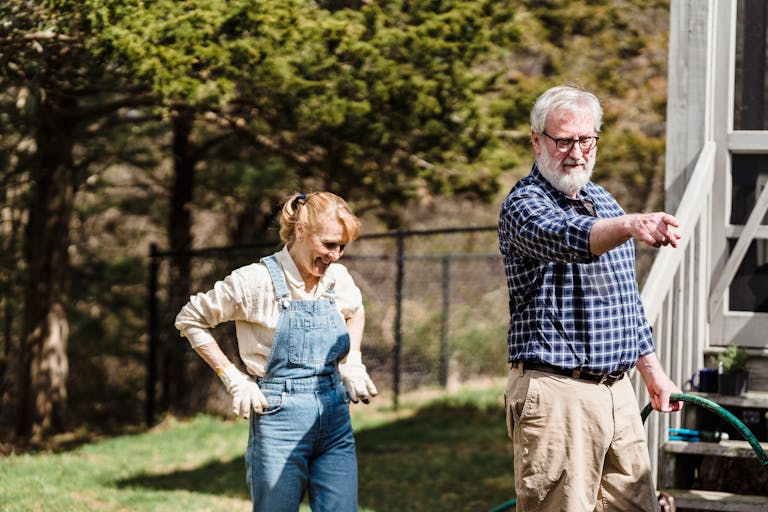 Couple of mature farmers with water hose standing on grassy lawn near building in countryside with green trees on summer day