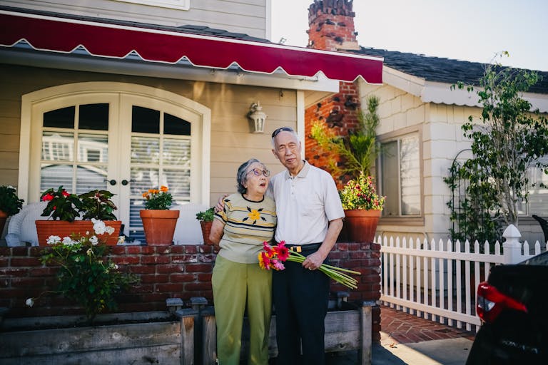 Elderly Asian couple embracing outside their cozy home with flowers, representing love and togetherness.