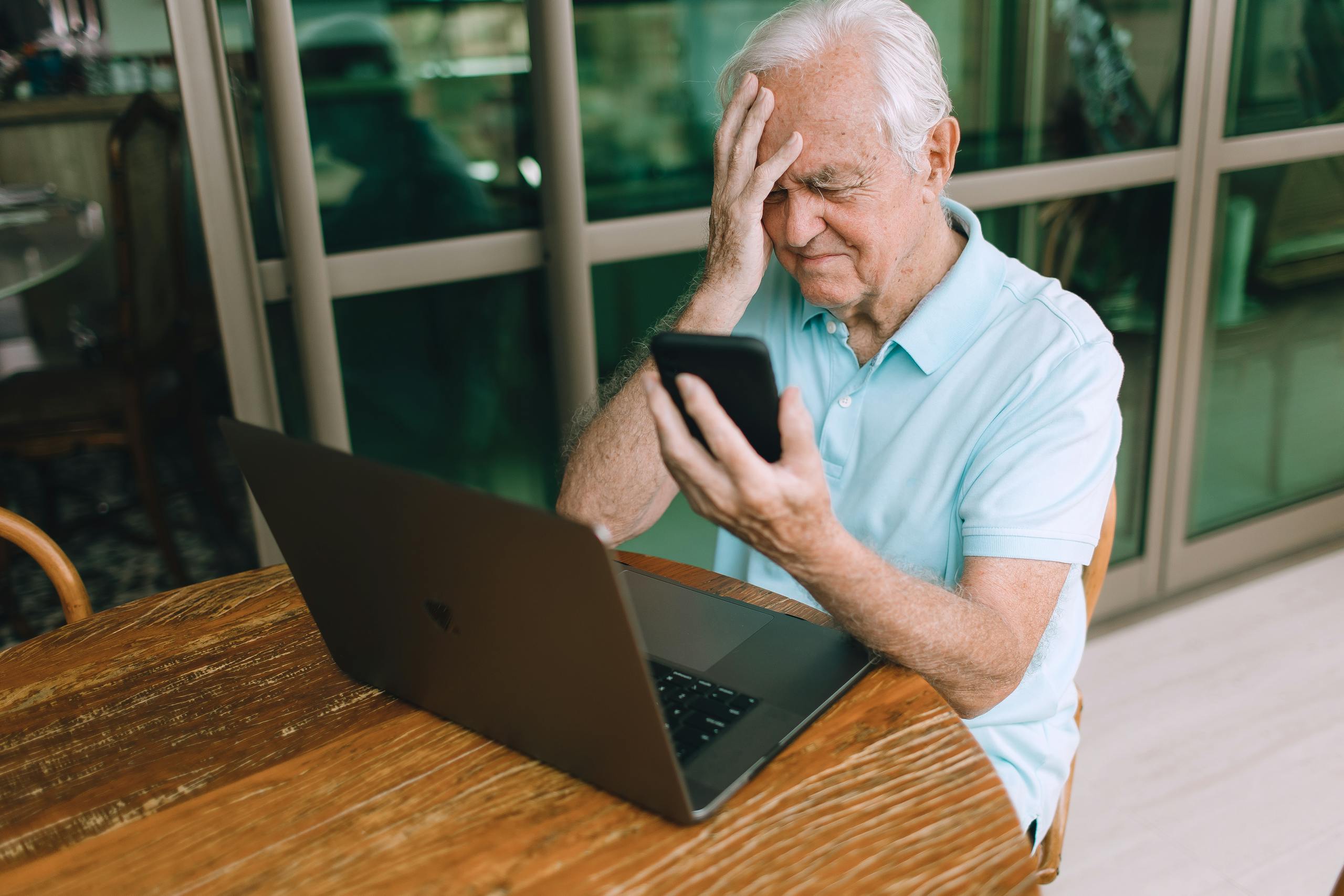Elderly man frustrated by smartphone and laptop technology indoors at home.