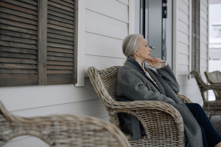 Elderly woman sitting in a cozy wicker chair on a porch, reflecting in winter wear.