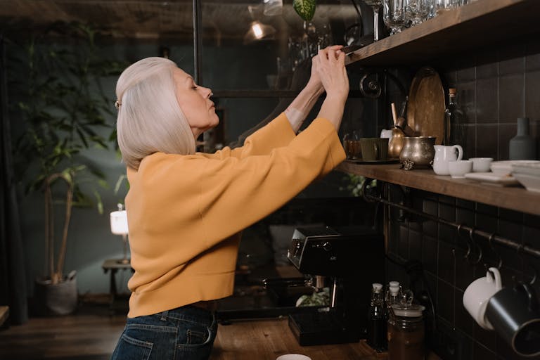 Elderly woman with white hair reaching a kitchen shelf in a cozy kitchen setting.