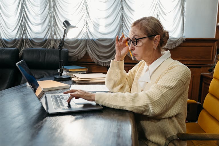 Elderly woman working on a laptop in an office, wearing eyeglasses and a yellow sweater.