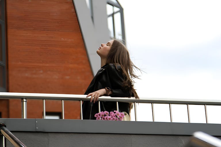 Elegant woman enjoying outdoors on an urban balcony, holding pink flowers.
