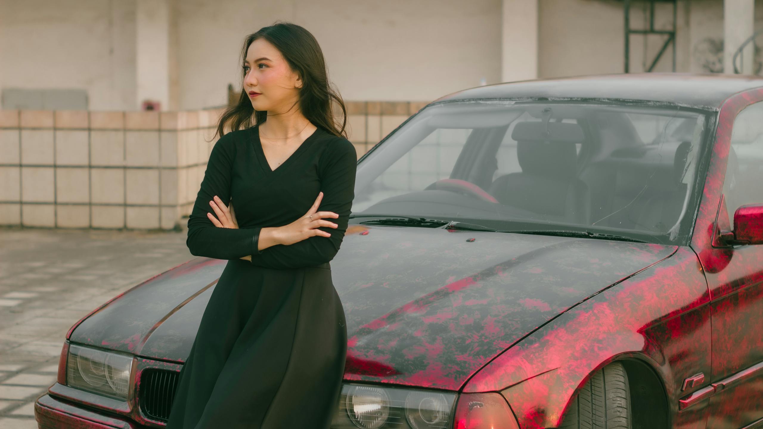 Fashionable woman in black attire posing confidently by a red car in an outdoor setting.
