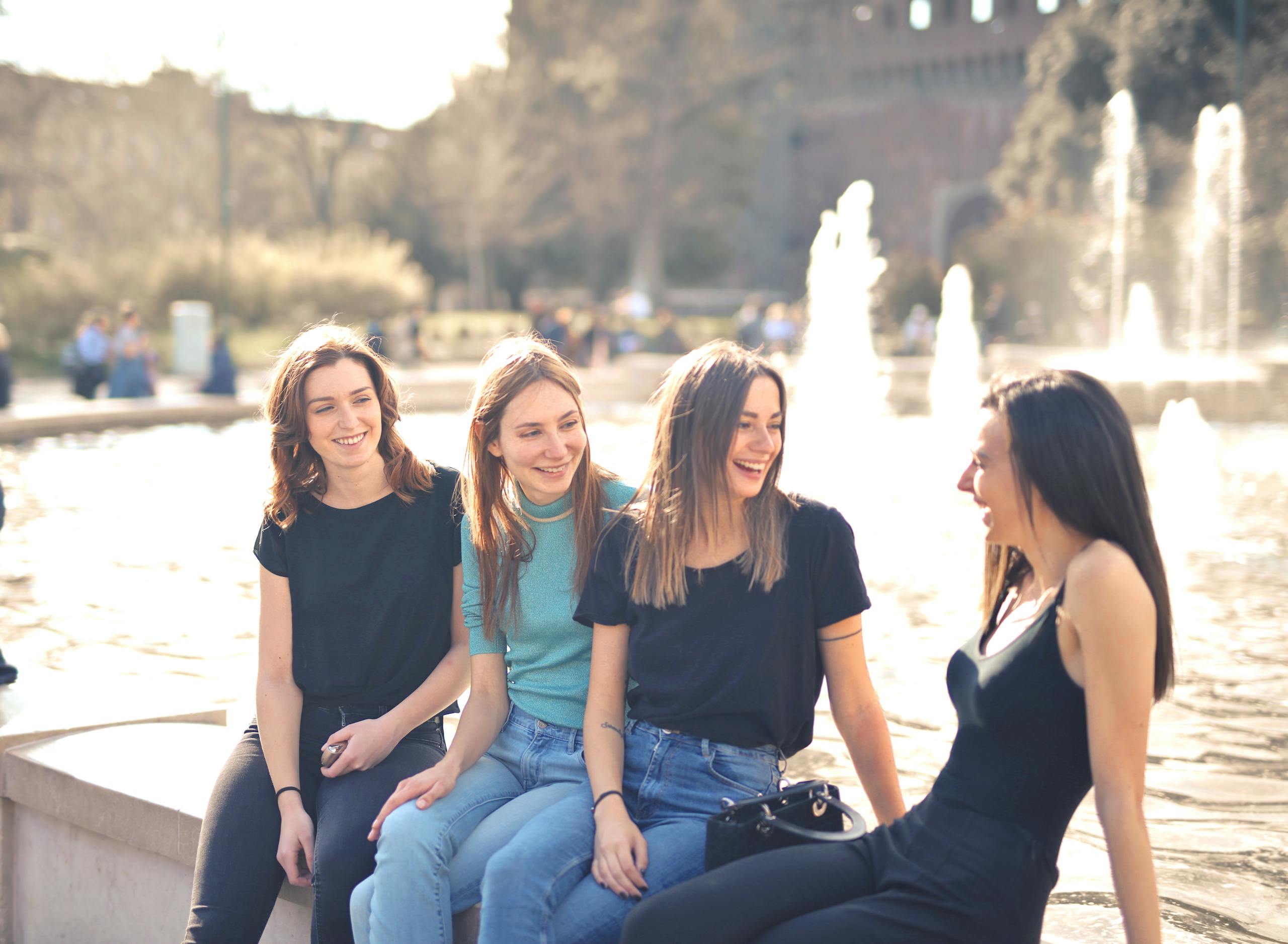 Four friends laughing and chatting outdoors by a water fountain on a sunny day.