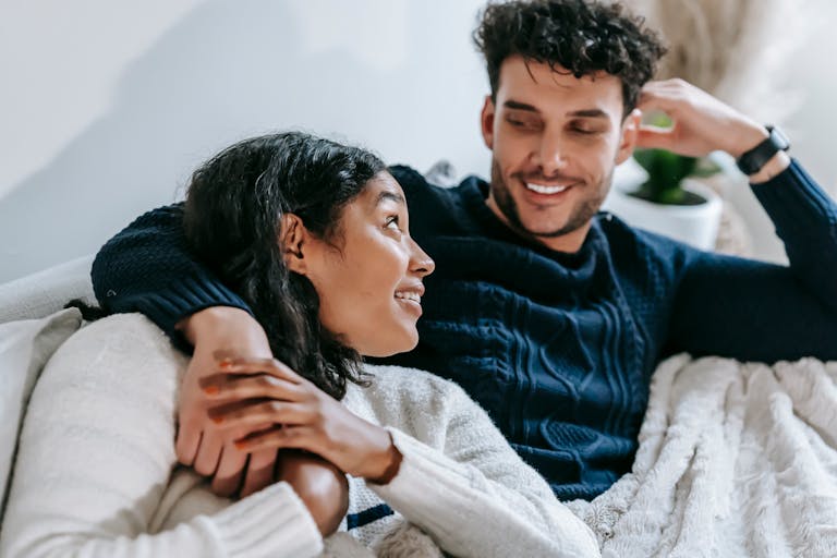 From above of positive African American girlfriend and ethnic boyfriend hugging and looking at each other while sitting on sofa under plaid