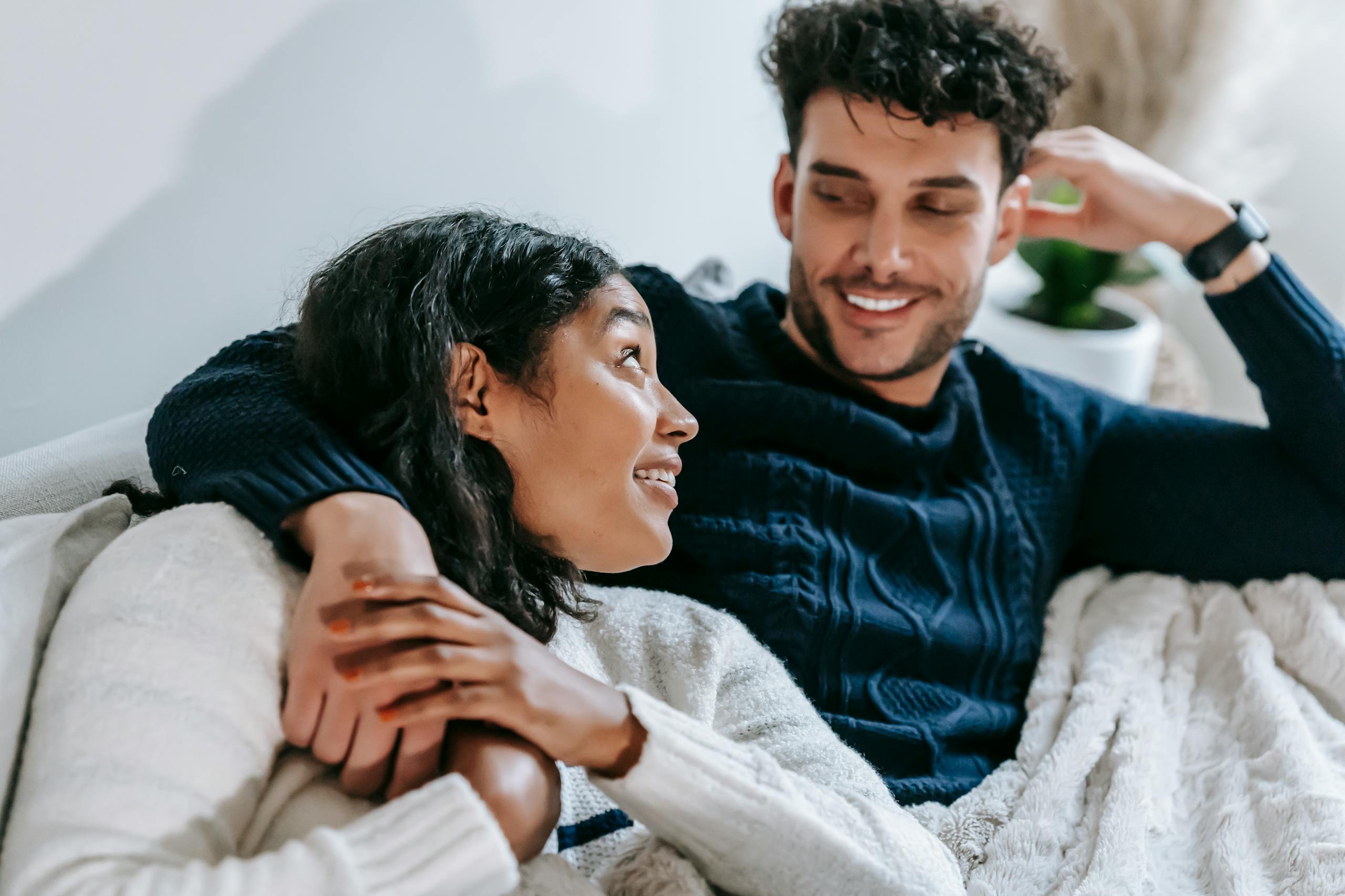From above of positive African American girlfriend and ethnic boyfriend hugging and looking at each other while sitting on sofa under plaid
