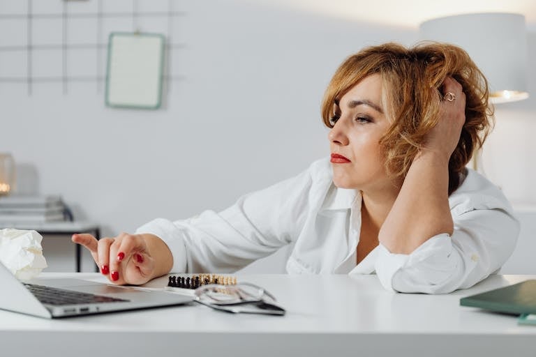 Frustrated woman at desk, using laptop with expression of stress. Indoors office environment.