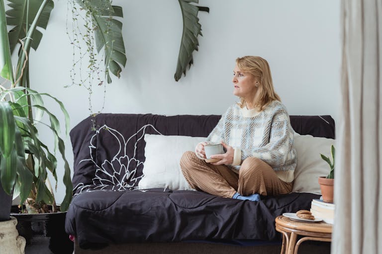 Full body of peaceful middle aged woman with blond hair in casual clothes drinking coffee and looking away thoughtfully while resting on sofa with crossed legs