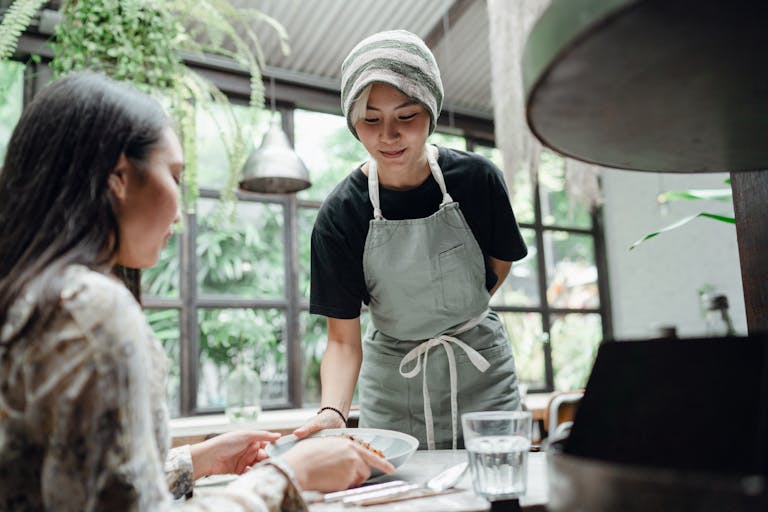 Glad young Asian waitress in uniform serving plate with tasty dish to female customer sitting at table in modern light restaurant against big window and plants