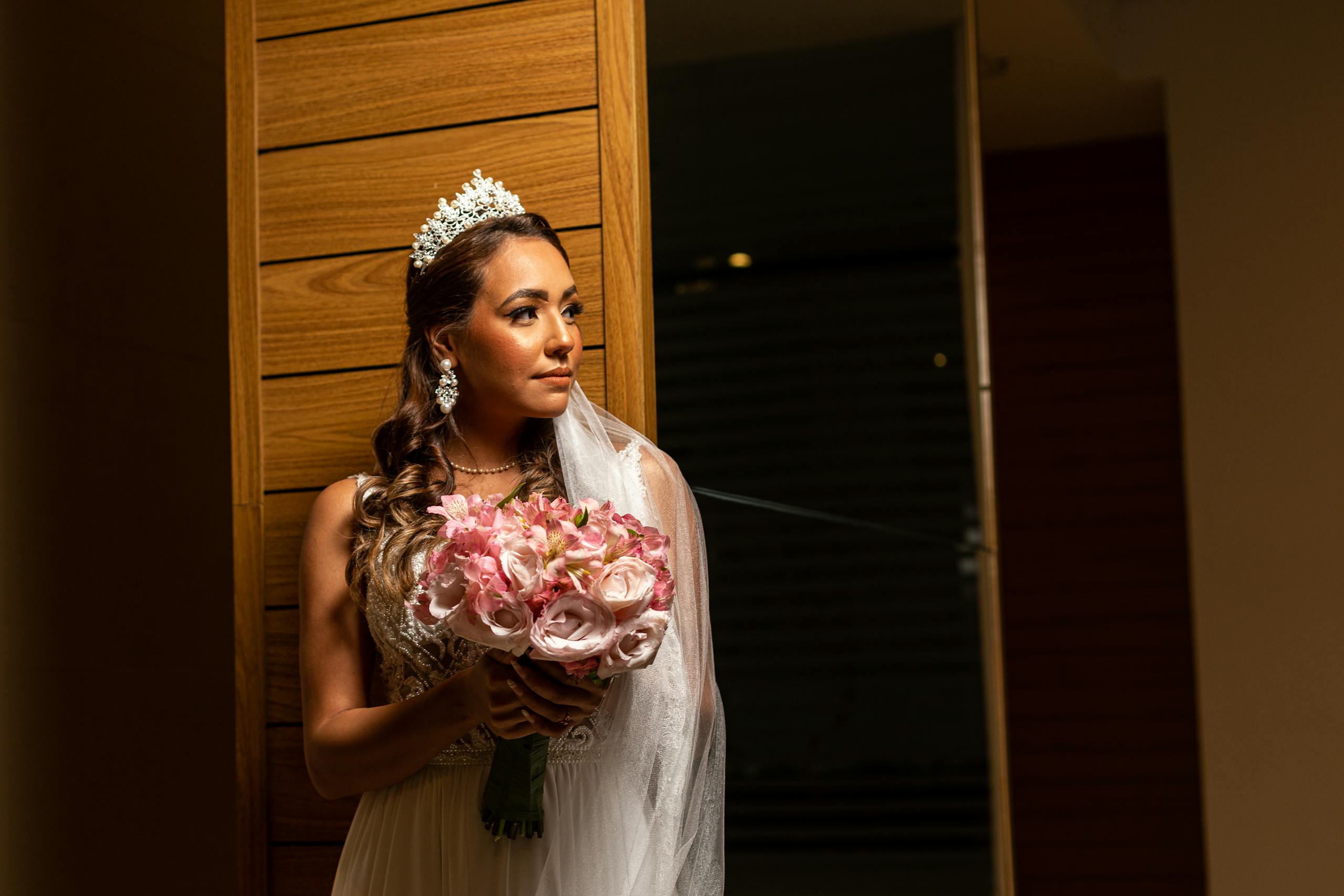 Glamorous bride in white dress holding a pink bouquet, looking afar indoors.