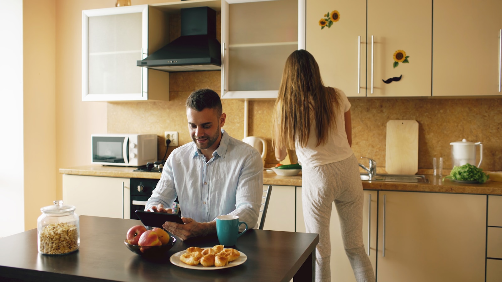 Couple in kitchen, man using tablet, woman preparing food.