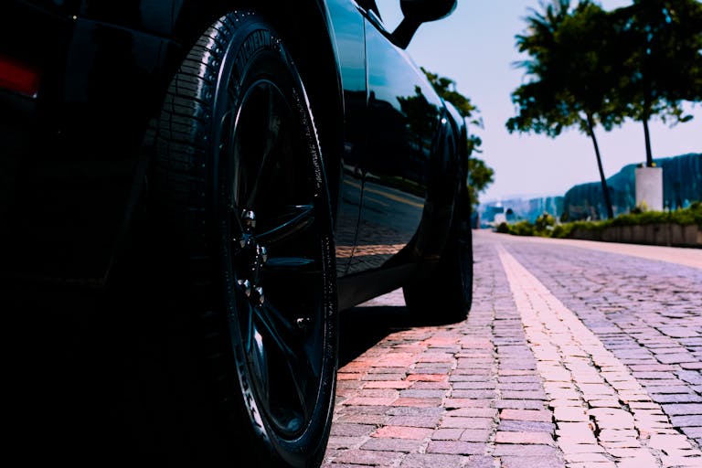 Low-angle view of a black car on a sunny cobblestone street lined with trees, perfect for urban lifestyle themes.
