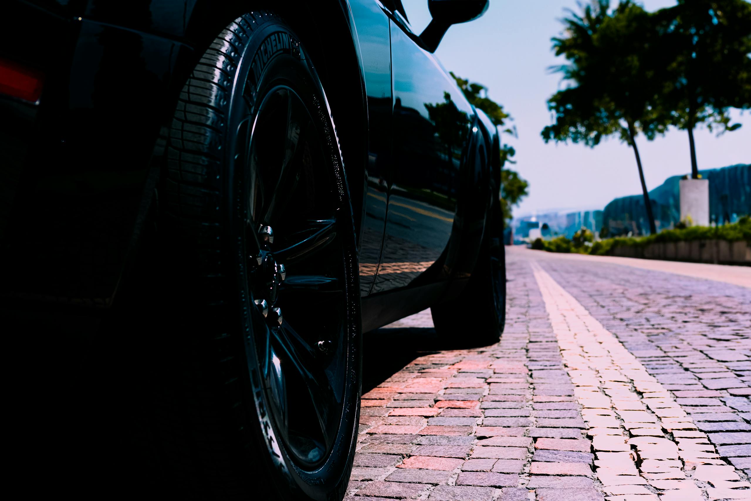 Low-angle view of a black car on a sunny cobblestone street lined with trees, perfect for urban lifestyle themes.