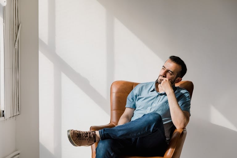 Man in casual attire sitting thoughtfully in a sunlit room, deep in contemplation.