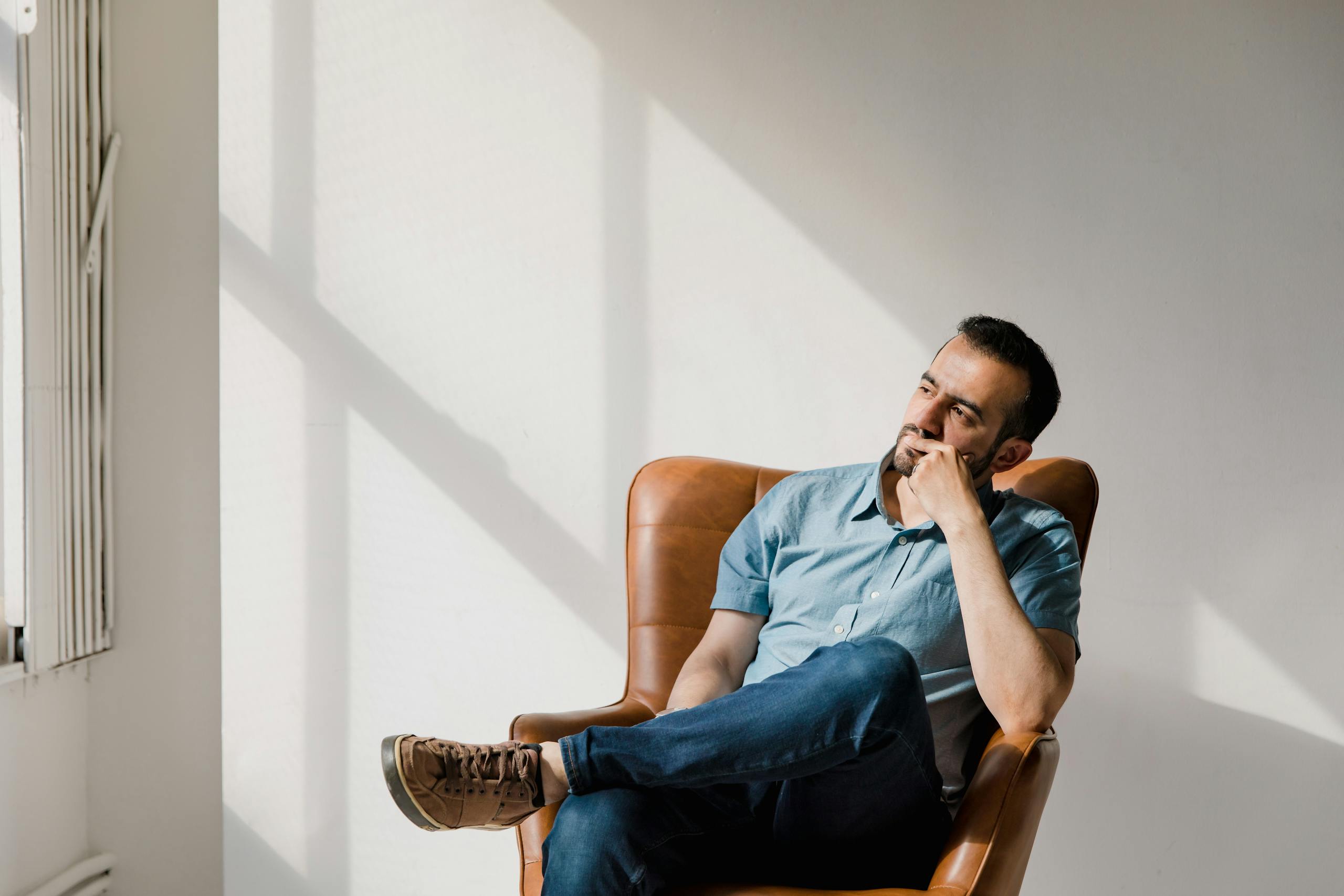 Man in casual attire sitting thoughtfully in a sunlit room, deep in contemplation.