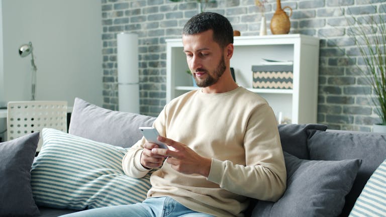 Man sitting on a couch using a smartphone in a cozy living room setting.