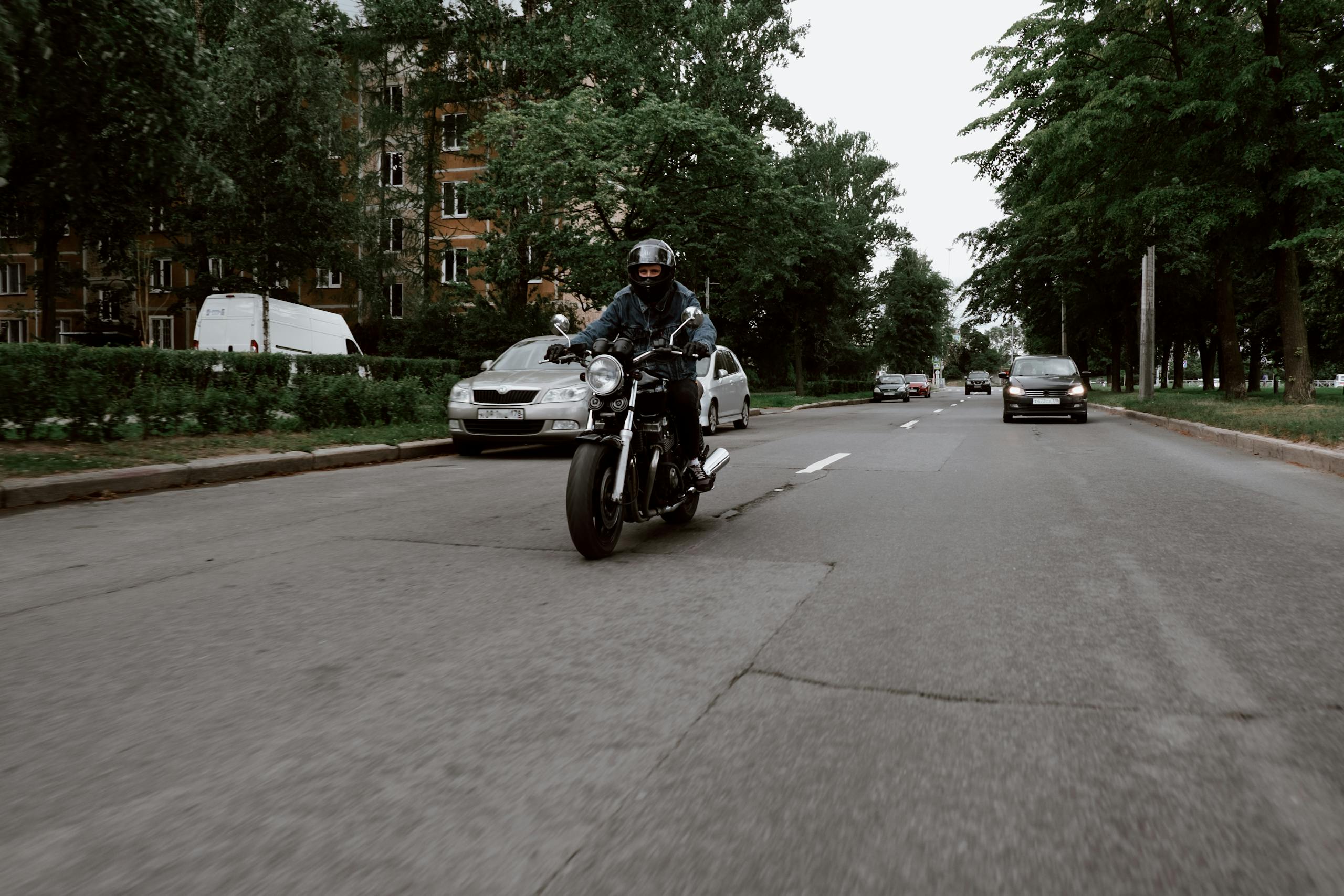Motorcyclist wearing denim and helmet riding on a tranquil road flanked by trees and vehicles.