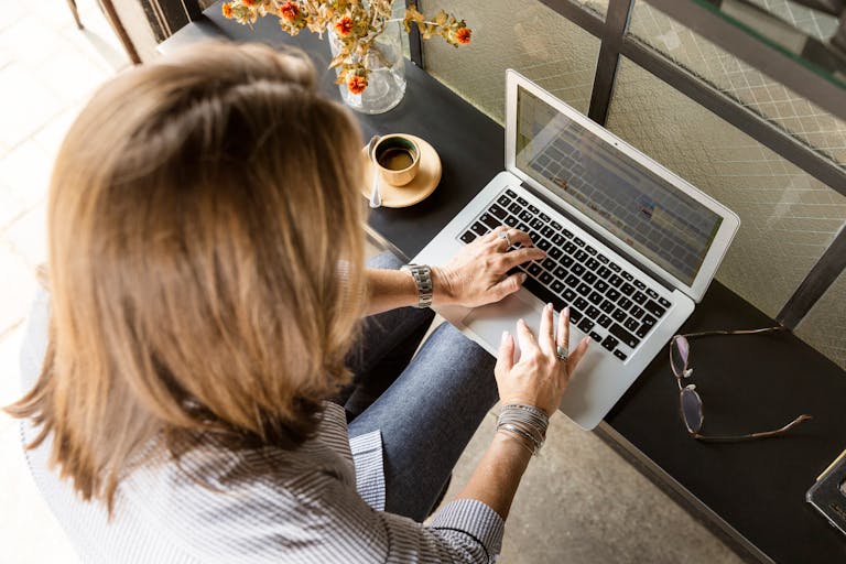 Overhead view of a woman typing on a laptop at a desk with coffee and flowers indoors.