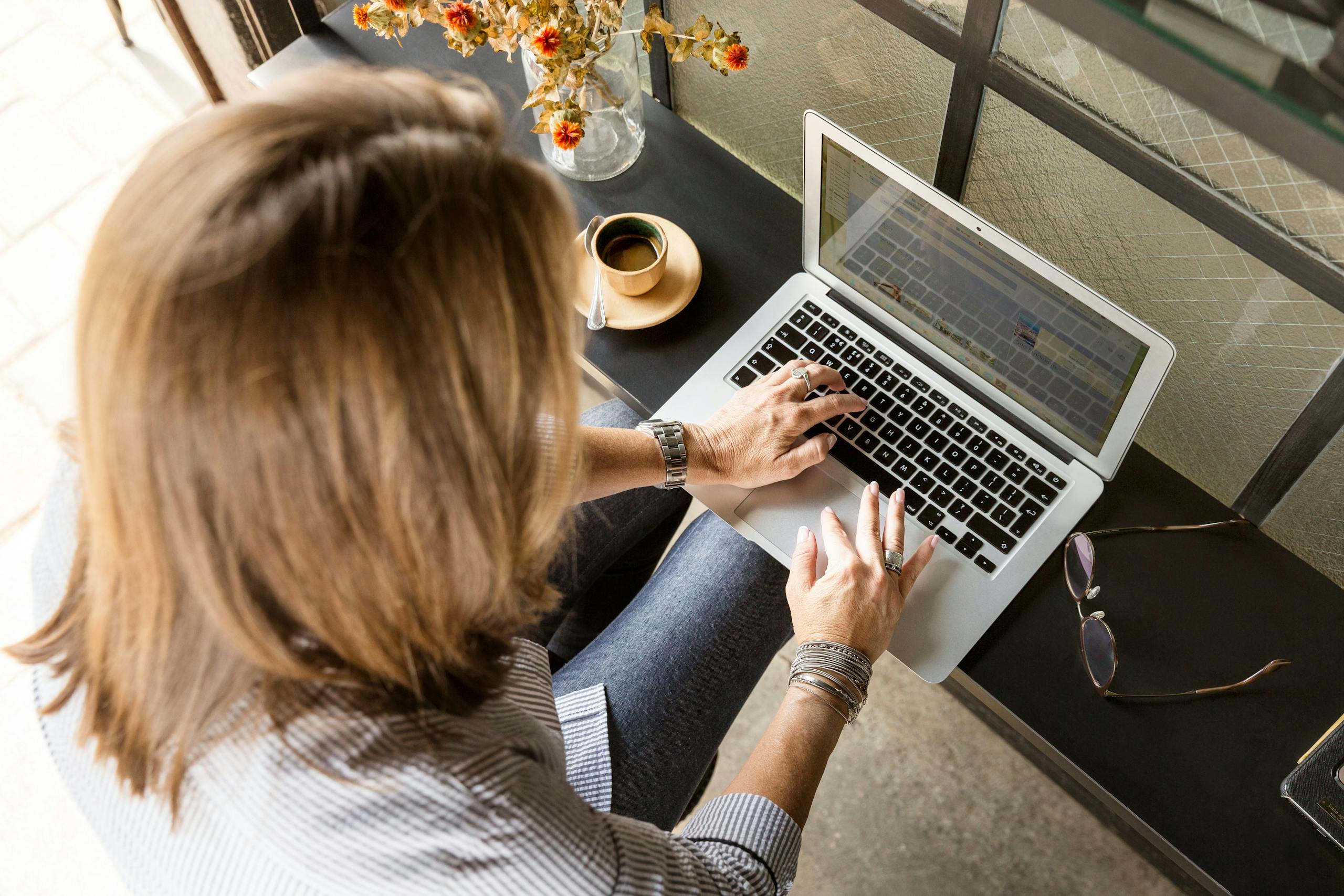 Overhead view of a woman typing on a laptop at a desk with coffee and flowers indoors.