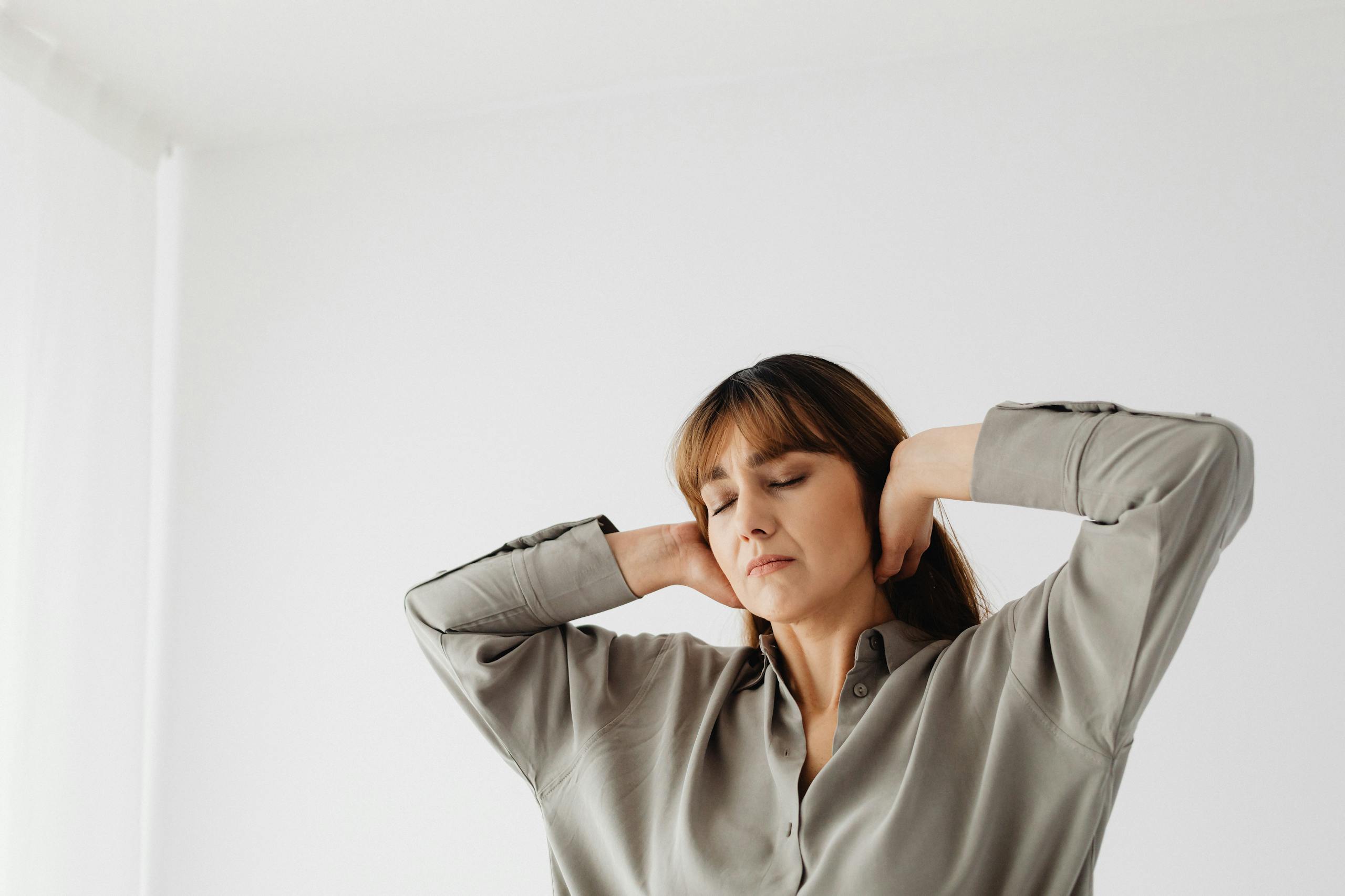 Portrait of a relaxed woman with eyes closed and hands behind head, in a serene indoor setting with white background.