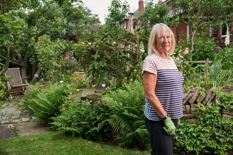 Positive adult female in casual clothes with rubber gloves and tools while working in greenery blooming garden