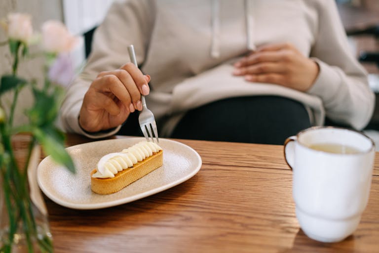 Pregnant woman in hoodie savoring a dessert with tea at a cozy cafe setting.