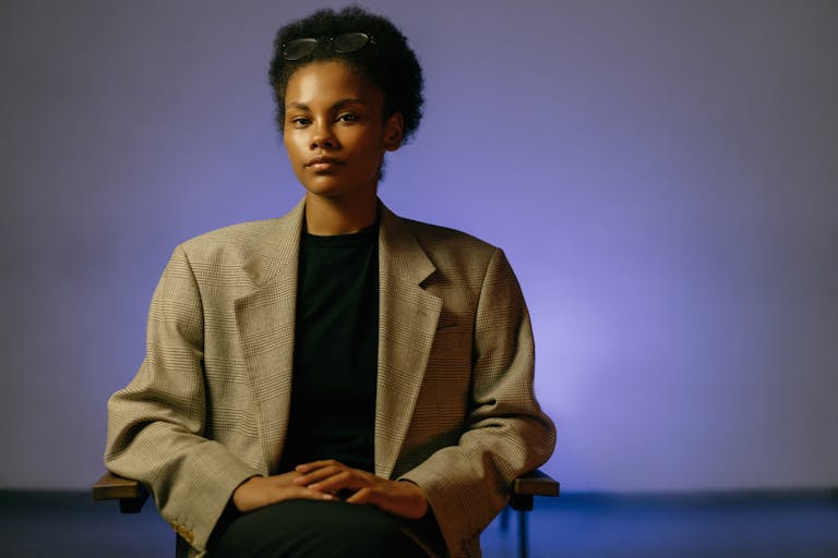 Professional woman with curly hair and eyeglasses sitting confidently in a studio.