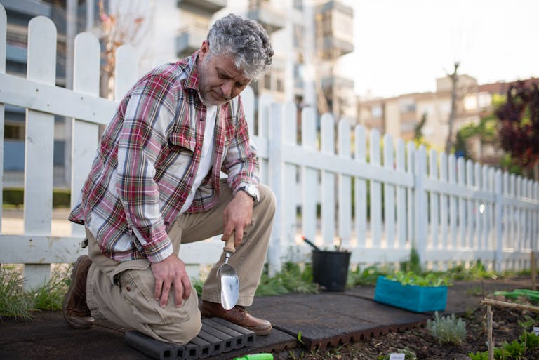 Senior man gardening with a trowel near a white fence in an urban garden, enjoying leisure time.