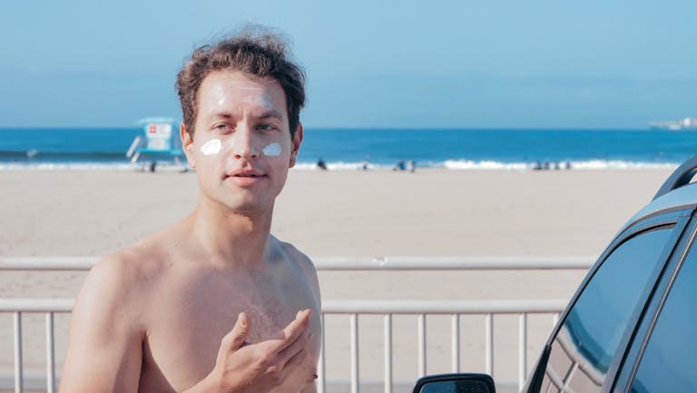 Shirtless man at the beach with sunscreen cream on his face, enjoying a sunny day.
