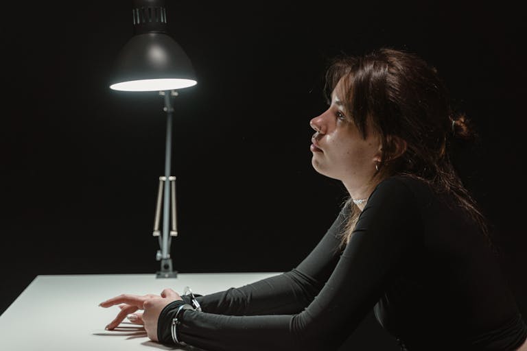 Side view of a woman sitting in an interrogation room under a lamp, handcuffed at desk.