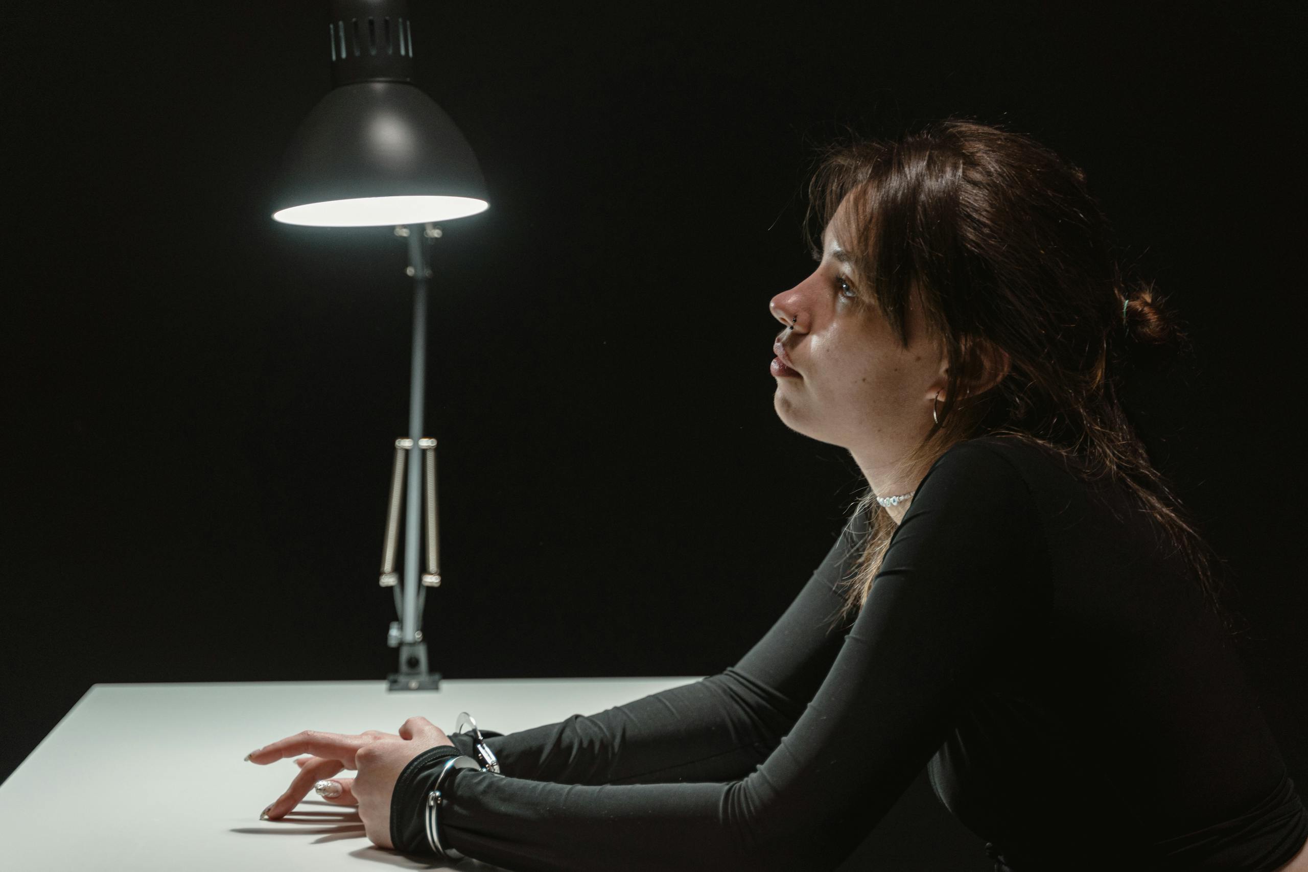 Side view of a woman sitting in an interrogation room under a lamp, handcuffed at desk.