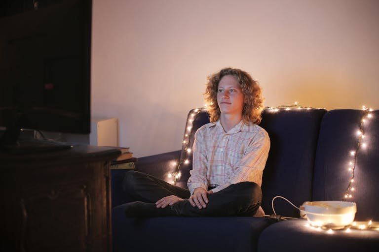 Smiling male wearing casual shirt sitting with crossed legs on cozy sofa and enjoying TV series while resting in dark room with garland during weekend