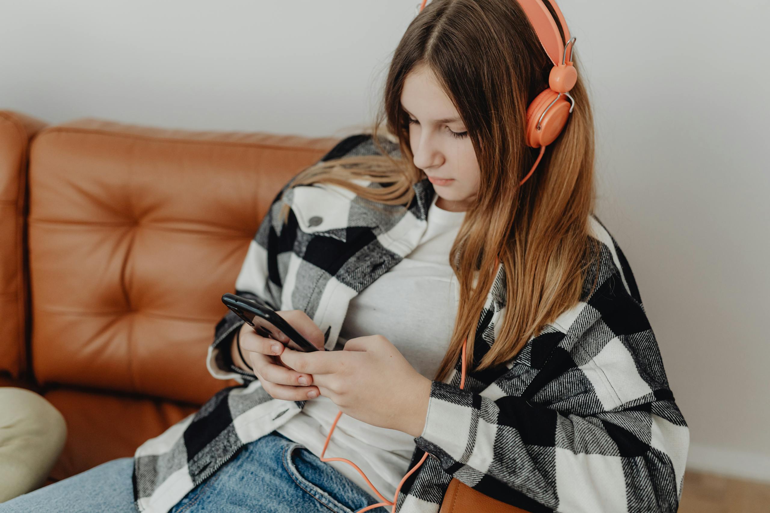 Teen girl sitting on a couch, listening to music with orange headphones, and using a smartphone.