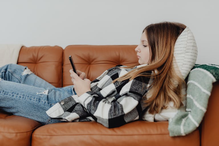 Teenager relaxing on a brown sofa using a smartphone, cozy and casual setting.