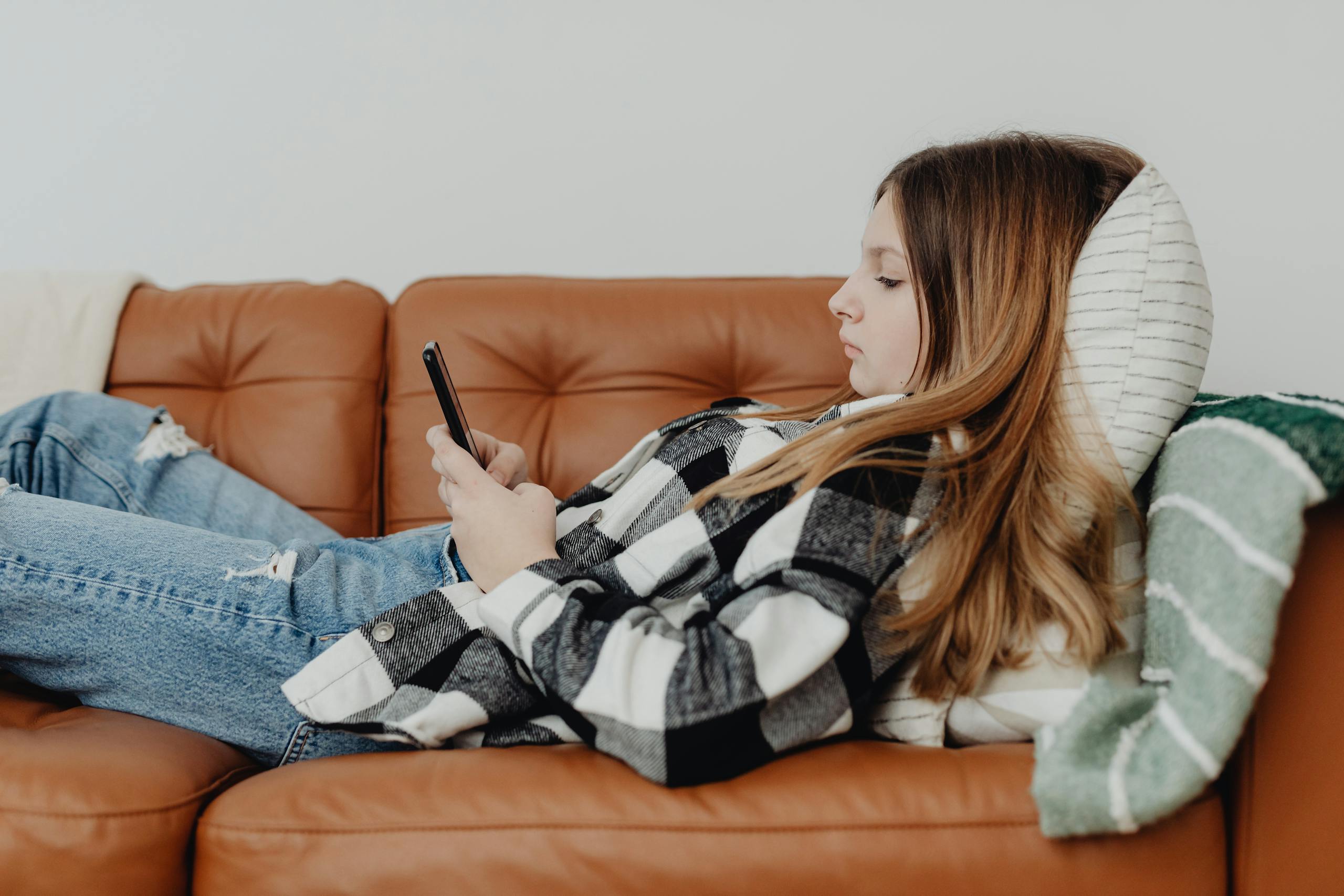 Teenager relaxing on a brown sofa using a smartphone, cozy and casual setting.