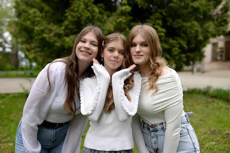Three teenage girls in casual outfits smile while posing together outdoors in a park setting.