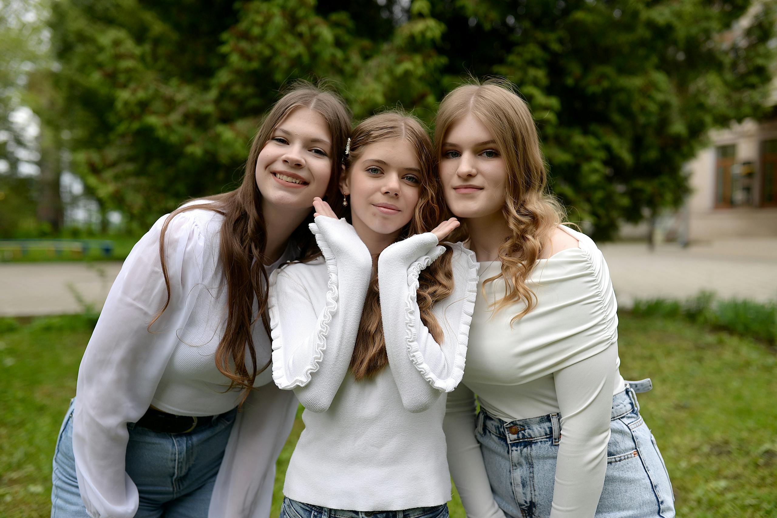 Three teenage girls in casual outfits smile while posing together outdoors in a park setting.
