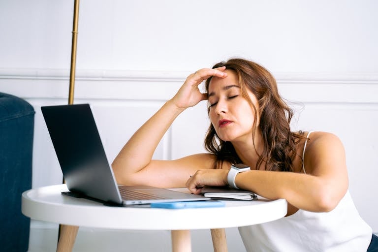 Tired woman with eyes closed, resting on a table with a laptop and smartwatch.