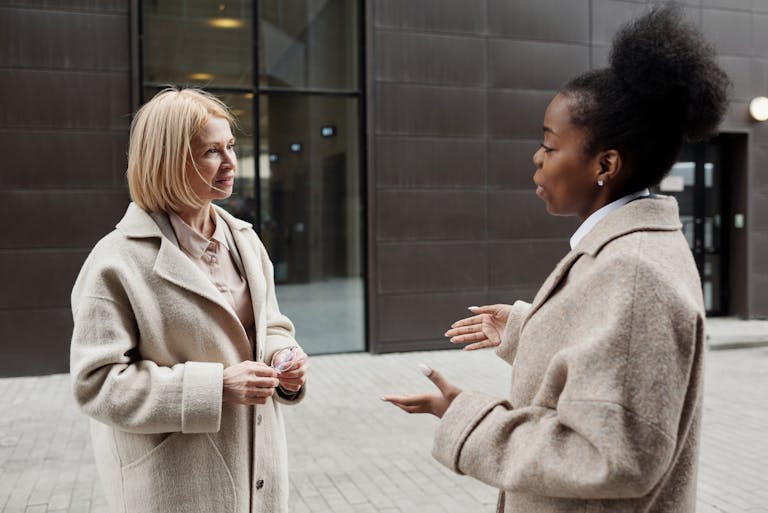 Two businesswomen in coats converse outside a modern office building.